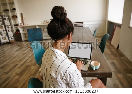 Back view of young curly woman with bun hairstyle in casual striped shirt typing text on keyboard and looking at screen, having cup of coffee on table