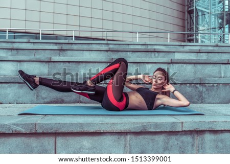 Girl in the summer in city on a yoga mat does exercises on the abs, twisting the abs. Active movement, sports figure and clothes, top leggings, sneakers.