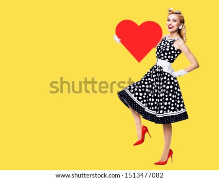 Full body of happy woman holding red heart symbol, dressed in pin up style black dress with white polka dot, isolated over yellow color background. Love, sales or retro fashion and vintage concept.