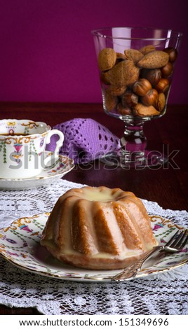 Mini Pound Cake - Almond lemon drizzle on old pictures coffee cup, side plate on lace and glass full of almonds and hazelnuts, purple background