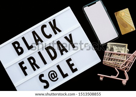 Creative promotion composition Black friday sale text on lightbox on black background, next grocery trolley, mobile phone, credit card, cash money. Flat lay, top view, overhead, mockup