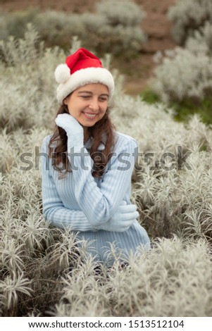 Beautiful young woman wearing red santa hat. Happy Christmas and New Year. Pretty Caucasian woman with smile looking down and touching her cheek with hand in gloves. Winter season. Edelweiss valley.