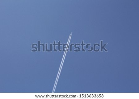 Vertical photo of a large white plane. A plane flies through the sky. The sky is blue. After the plane, a trace is visible.