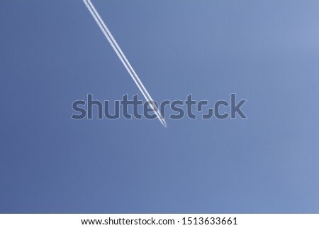 Vertical photo of a large white plane. A plane flies through the sky. The sky is blue. After the plane, a trace is visible.