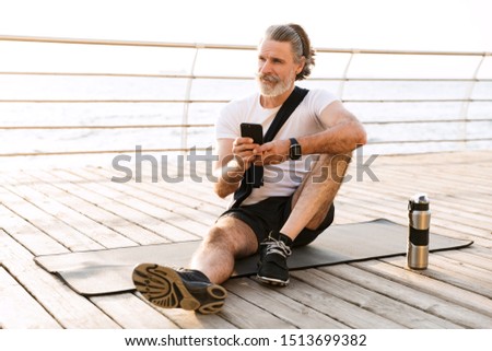 Image of pleased elderly man in sportswear typing on cellphone while sitting on mat near seaside in morning