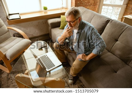 Senior man working with laptop at home - concept of home studying. Caucasian male model sitting on sofa and doing his homework or serfing in internet, watching cinema or webinar, playing games.