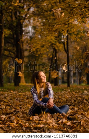 Beautiful smiling girl sitting on the ground on yellow leaves in park. Leisure time on warm autumn day, sunset light