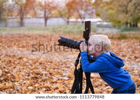 Little blond boy shoots with a large SLR camera on a tripod. Photo session in the autumn park