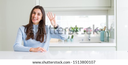 Wide angle picture of beautiful young woman sitting on white table at home showing and pointing up with fingers number four while smiling confident and happy.