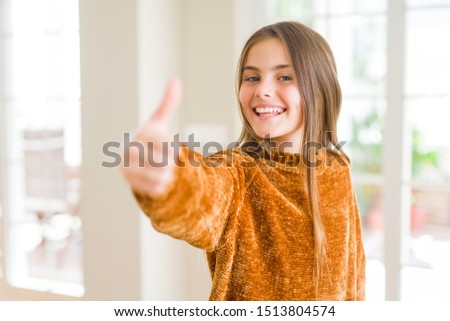 Beautiful young girl kid at home doing happy thumbs up gesture with hand. Approving expression looking at the camera showing success.