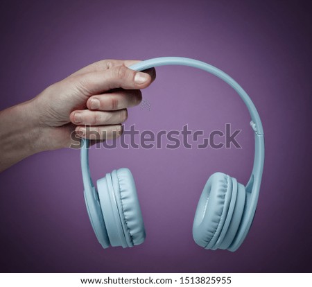 Female hand holding modern wireless blue headphones on a purple background.