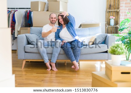 Young couple sitting on the sofa arround cardboard boxes moving to a new house smiling making frame with hands and fingers with happy face. Creativity and photography concept.