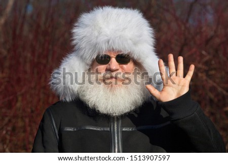 Smiling senior caucasian man in white fur hat and sunglasses showing hi gesture. Closeup portrait of elderly stylish hipster in black flight (bomber) jacket against dark forest background.