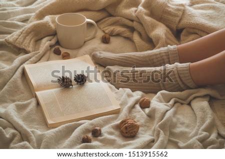 Cozy Autumn winter evening , warm woolen socks. Woman is lying feet up on white shaggy blanket and reading book. Cozy leisure scene. Text in book is unreadable. Woman relaxing at home. Comfy lifestyle