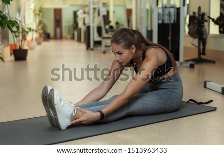 Young attractive fit woman doing leg stretching exercise while sitting on mat in gym