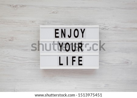 'Enjoy your life' words on a lightbox over white wooden background, top view. Overhead, from above. Flat lay. 