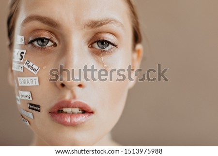 Blue-eyed lady with words on her face is crying, looking into camera on beige background