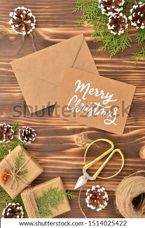 Brown kraft envelope with calligraphy handwritten sign Merry Christmas on wooden table with vintage tone. Top view New Year frame with pine cones, gift boxes and fir branches