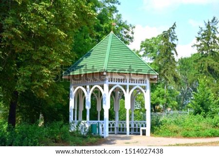 A large hut or shack with white wooden walls and arches and a slanted green roof made out of tiles standing in the middle of a public park on a summer day seen on a Polish countryside near a dirt road