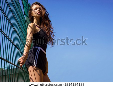 young girl with long brunette hair in sports black tank top and shorts is standing with closed eyes under the sun near grid on blue sky background, free space on right side