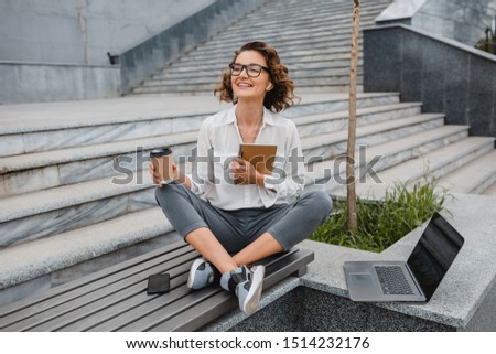 attractive stylish smiling woman in glasses working typing on laptop, freelancer sitting in park on stairs drinking coffee, urban city street style, business center, online education