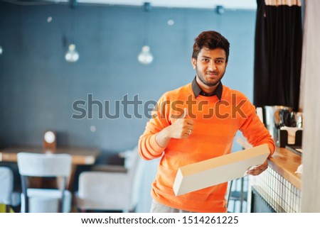 Confident young indian man in orange sweater standing near bar counter at cafe and hold box for delivery with pizza.