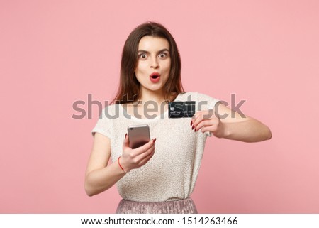 Shocked young woman in casual light clothes posing isolated on pastel pink background. People lifestyle concept. Mock up copy space. Using mobile phone, typing sms message, holding credit bank card