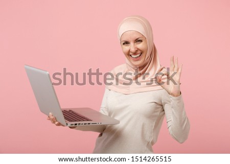 Joyful young arabian muslim woman in hijab light clothes posing isolated on pink background. People religious lifestyle concept. Mock up copy space. Hold, work laptop pc computer, showing OK gesture