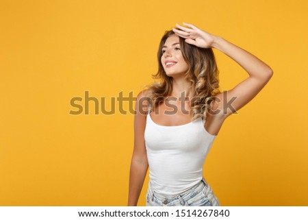 Smiling young woman girl in light casual clothes posing isolated on yellow orange wall background. People lifestyle concept. Mock up copy space. Holding hand at forehead looking far away distance
