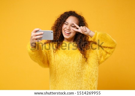 Cheerful young african american girl in fur sweater posing isolated on yellow orange background. People lifestyle concept. Mock up copy space. Doing selfie shot on mobile phone, showing victory sign