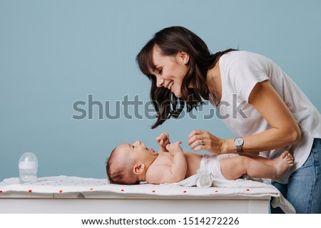 Mother changing diaper on her baby on table over blue background. They look at each other, smile and laugh. Royalty-Free Stock Photo #1514272226