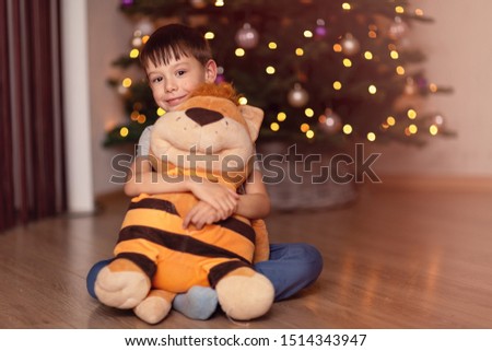 A charming five year old boy sits with a large plush toy on the floor amid the glowing lights of a Christmas garland. Cute baby hugs from behind the neck of his toy friend and smiles.