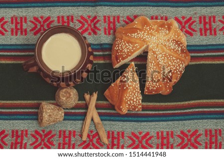 Pan de muerto and cup of chocolate on a table with typical Mexican tablecloth. With preparation ingredients such as cinnamon and brown sugar