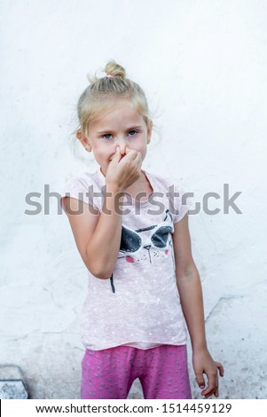 little blonde girl in pink stands at the white stele and covered her nose with fingers