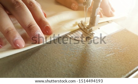 A woman worker is sewing the clothes for making a dress with light flare on the right corner of the picture. 