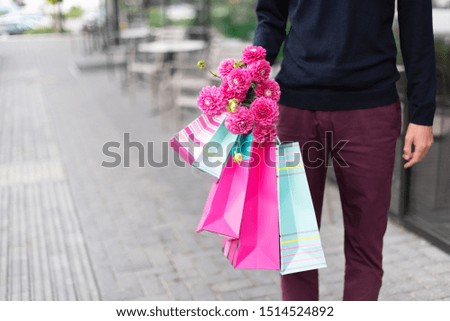 Stylish man holding flowers and pink, blue shopping bags near restaurant. Woman's day. Valentine's day. Ready for birthday party or romantic date. Copy space. Bunch of paper gift bags.