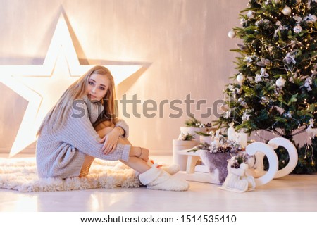 Beautiful young blond woman dressed in white sweater posing under Christmas tree in a holiday interior
