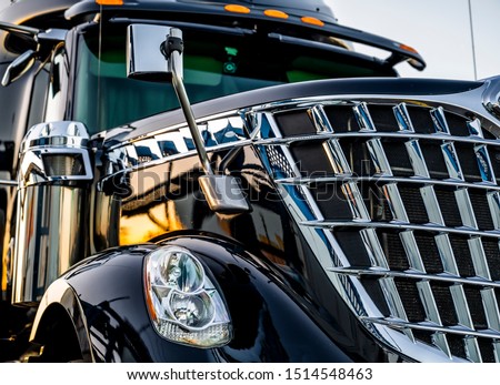 Big rig classic American idol black industrial semi truck with chrome grille and commercial cargo in dry van semi trailer standing on the truck stop parking lot at twilight time reflection sunlight