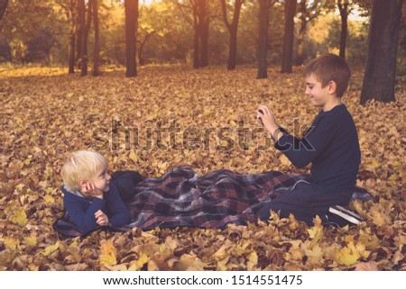 Two little brothers take pictures of each other, lying in yellow autumn leaves. Fall day