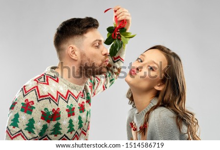 christmas, people and holiday traditions concept - portrait of happy couple in ugly sweaters kissing under the mistletoe over grey background
