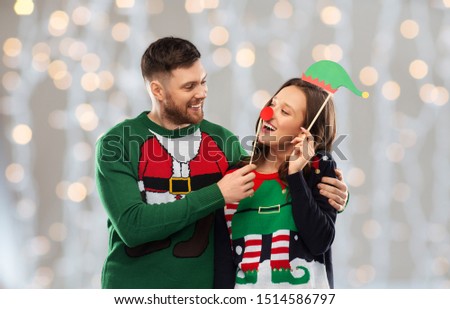 christmas, photo booth and holidays concept - happy couple in ugly sweaters posing with party props over festive lights background