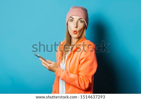 Beautiful Young woman in a hoodie and a hat with a surprised face holds a mobile phone on a blue background. Concept winter, christmas, winter holidays, application for the phone.