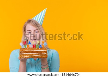 Portrait of a funny positive girl with a paper cap holding a congratulatory cake in her hands on a yellow background with copy space. Concept and fun and celebration. Advertising space.
