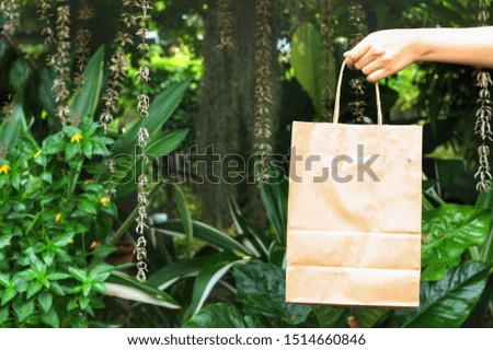 Female hand is holding paper shopping bag with blurred green forest background.Using Paper container for takeaway food or things can reduce plastic bag.Ecological,reusable concept.Have copy space.