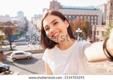 Happy beautiful woman smiling, taking selfies in city center. Gorgeous cheerful young woman enjoying wandering in the city, taking a selfie