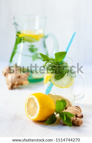 Healthy and tasty drink: homemade lemonade with lemon, mint, ginger in transparent glass mug with blue straw. Healthcare, concious lifestyle, detox. White background, jug in the background