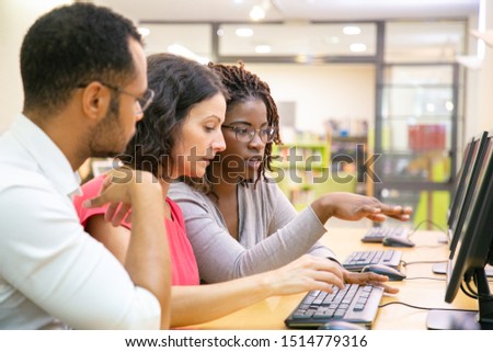 Instructor explaining corporate software specific to trainees in computer class. Man and women sitting at table, using desktop, pointing at monitor and talking. Training concept Royalty-Free Stock Photo #1514779316