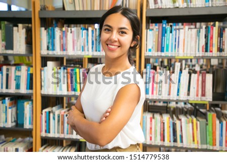 Smiling Asian woman posing at public library. Front view of smiling lady with crossed arms posing in front of bookshelves. Knowledge concept