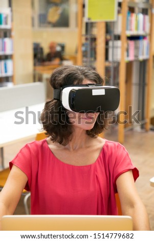 Happy adult student using VR headset during work in library. Middle aged woman in virtual reality glasses sitting at desk with laptop, headset turning face up and smiling. Education concept