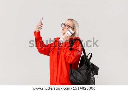 Blond girl in a red raincoat and glasses making selfie with a smartphone travelling with black backpack and white thermal mug in cold season. Rainy season.